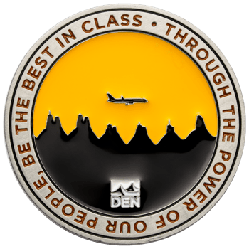 Round challenge coin featuring the DEN logo, a black mountain skyline, and a silhouette of an airplane against a yellow sky.